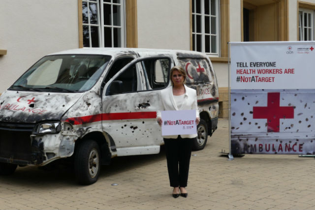 Luxembourg Grand Duchess Maria Teresa stands in front of a damaged ambulance holding a sign that reads "#NOTATARGET" Luxembourg Red Cross