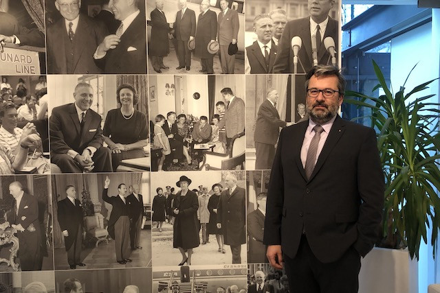 Ambassador Bärtl standing before a collage of historical images at the Czech embassy in Limpertsberg Delano